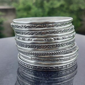 Puede incluir: Una pila de pulseras plateadas con intrincados diseños florales y geométricos. Las pulseras están apiladas, mostrando los diseños detallados y la superficie reflectante del metal. Las joyas se exhiben sobre una superficie oscura.