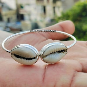 Op de afbeelding: Een zilveren armband met twee grote, natuurlijke kaurischelpen. De schelpen zijn crèmekleurig en gezet in zilver metaal. De armband wordt op een hand getoond, met de tekst "silverartisanitem" zichtbaar.