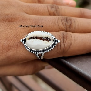 May include: A silver ring with a white cowrie shell set in a beaded oval frame. The ring is on a finger.