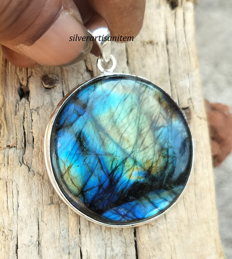 May include: Round pendant with a vibrant, iridescent labradorite stone. The stone displays shades of blue, green, and black, set in a polished silver bezel. The pendant has a silver loop for a chain.