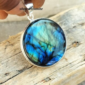 May include: A round pendant with a vibrant blue and green gemstone, set in a silver-colored metal frame. The gemstone displays a unique pattern of colors. The pendant is held by a hand, and the text "silverartisanite" is visible.