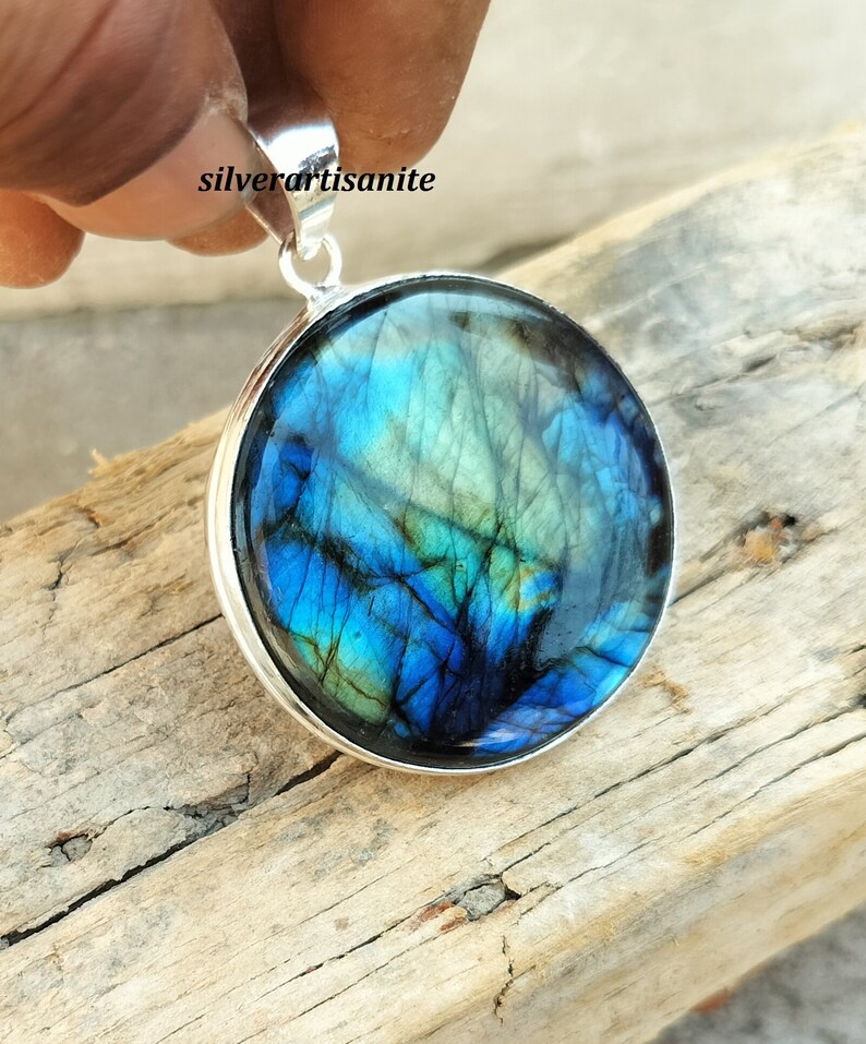 May include: A round pendant with a vibrant blue and green gemstone, set in a silver-colored metal frame. The gemstone displays a unique pattern of colors. The pendant is held by a hand, and the text "silverartisanite" is visible.