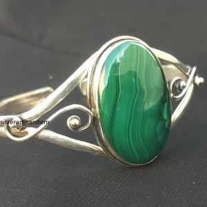 May include: Silver cuff bracelet with a large, oval malachite stone. The stone displays green and black patterns. The bracelet has an open design with decorative swirls. The jewellery is displayed on a dark surface.