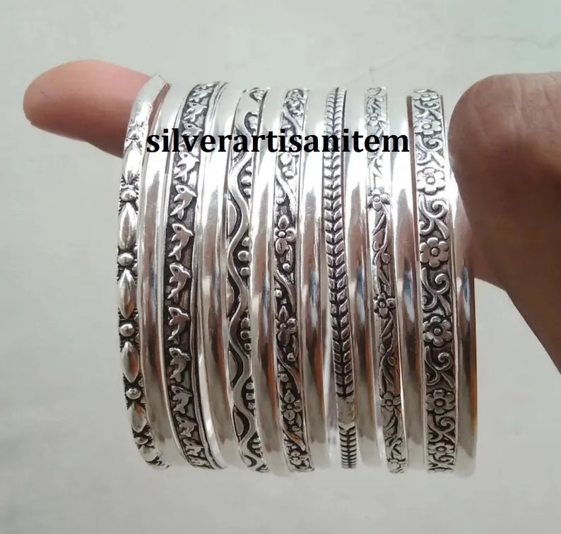 K&ouml;nnte beinhalten: Ein Set aus zehn silbernen Armreifen mit aufwendigen, ge&auml;tzten Designs. Die Armreifen sind &uuml;bereinander gestapelt und haben eine Vielzahl von Mustern, darunter florale, geometrische und abstrakte Designs.