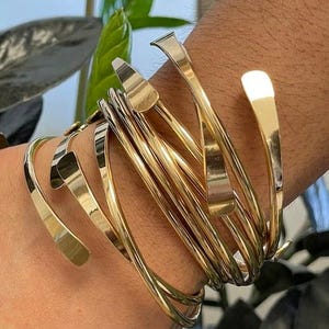 May include: A collection of gold-colored metal bracelets. The bracelets are of varying widths and designs, some with curved ends and others with a more angular shape. The bracelets are stacked on a wrist, creating a layered effect.