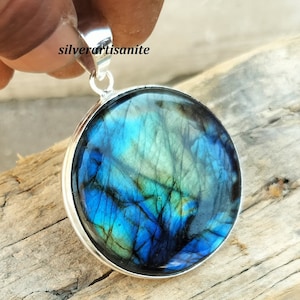 May include: Round pendant with a vibrant blue and green gemstone set in a silver bezel. The gemstone displays a unique pattern of colors. The pendant has a silver loop for a chain. The text "silverartisanite" is visible.