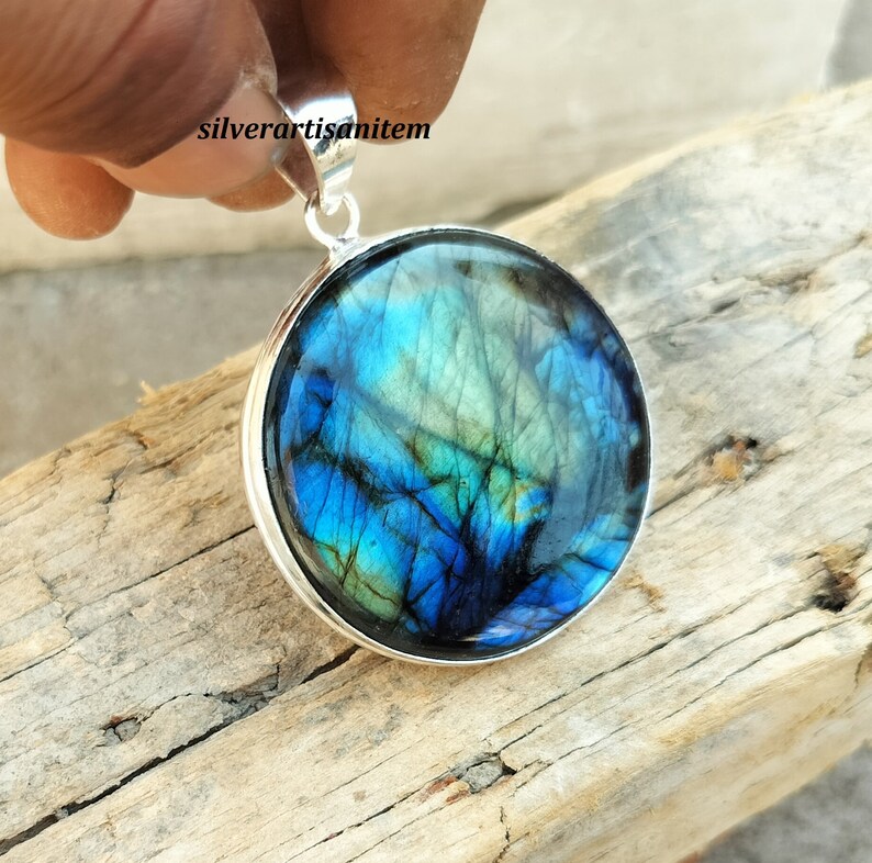 May include: Round pendant with a polished, iridescent labradorite stone, showcasing blue, green, and teal hues. The stone is set in a silver-colored metal bezel with a matching bail for a necklace chain. The text "silverartisanitem" is visible.