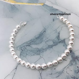 Può includere: Un braccialetto di perline d'argento con chiusura a moschettone. Il braccialetto è composto da perline rotonde e lucenti d'argento. Le perline sono infilate insieme per formare una catena flessibile. Il braccialetto è esposto su una superficie di marmo.
