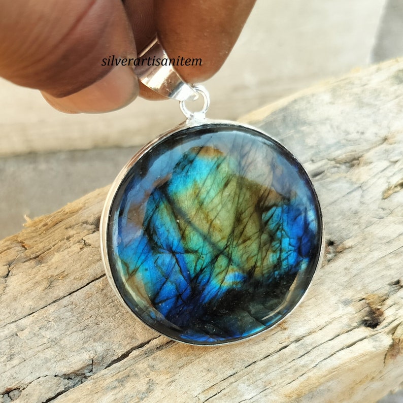 May include: Round pendant with a polished labradorite stone, showcasing iridescent blues, greens, and yellows. The stone is set in a silver-colored bezel with a matching bail for a necklace chain. The pendant is held up by a hand.