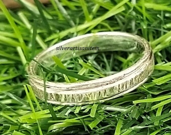 May include: A silver ring with a textured band and a pattern of small lines. The ring is lying on a bed of green grass.