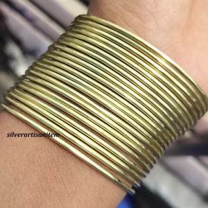 May include: A set of 20 gold-colored metal bangles stacked on a person's wrist. The bangles are thin and have a smooth, shiny finish.