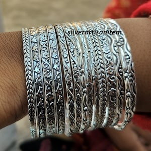 Pode incluir: Um conjunto de 12 pulseiras de prata com intrincados desenhos florais e geométricos. As pulseiras estão empilhadas no pulso de uma pessoa.