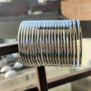 Puede incluir: Una pila de brazaletes de plata, un tipo de joyería tradicional india, están dispuestos en un patrón circular. Los brazaletes son lisos y brillantes, reflejando la luz.