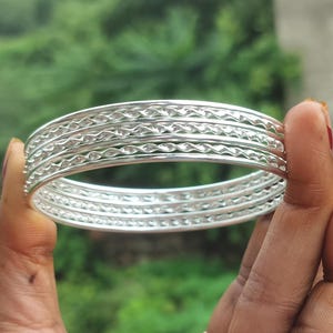 Puede incluir: Un conjunto de pulseras plateadas con intrincados patrones repetidos. Las pulseras se muestran sobre un fondo verde borroso, resaltando su superficie metálica reflectante. El diseño incluye bandas texturizadas y lisas.