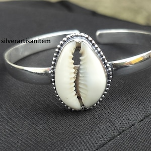 Può includere: Un braccialetto d'argento con una conchiglia di cauri bianca al centro. La conchiglia è circondata da un bordo di perline.