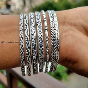 May include: A set of eight silver-toned bangles with various embossed floral and leaf designs. The bangles are stacked on a person's wrist.