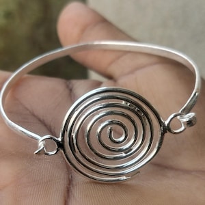Puede incluir: Un brazalete plateado con un diseño de espiral en el centro. El brazalete está hecho de un metal delgado y brillante, con un diseño simple y elegante. La espiral es el punto focal, creando una pieza de joyería única y llamativa.