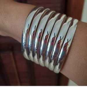 Op de afbeelding: Een stapel zilveren armbanden, gedragen op een pols. De armbanden zijn glad en glanzend en zijn op elkaar gestapeld.