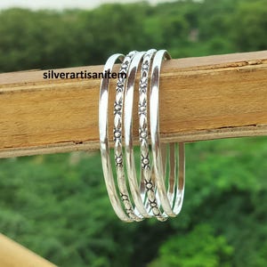 Op de afbeelding: Een set van zes zilveren armbanden met een bloemmotief. De armbanden worden getoond op een houten oppervlak tegen een vage groene achtergrond. De armbanden hebben een gepolijste afwerking en zijn even groot.