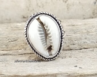 May include: A silver ring with a white seashell design. The ring has a rope-like border around the shell.