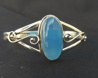 May include: A silver bangle bracelet featuring a light blue oval gemstone set in the centre. The bracelet has a delicate, ornate design with swirls on each side of the stone.