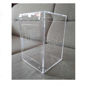 May include: A clear acrylic insect habitat with a small door and ventilation holes. The habitat is designed for small insects and has a square shape.