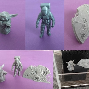 May include: Three 3D printed figurines: a grey astronaut, a grey Baby Yoda, and a grey, partially broken, circular object. The figurines are displayed on a purple surface and in a clear display case with a black base.