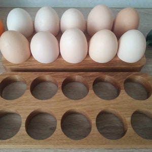 Könnte beinhalten: Ein hölzerner Eierhalter mit zehn kreisförmigen Aussparungen, der ein Dutzend Eier in verschiedenen Weiß- und hellbraunen Farbtönen enthält. Die Eier sind auf einer hellen Holzoberfläche angeordnet, mit einer Teilansicht einer Gurke und einer Orange.