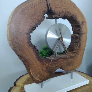 May include: A distinctive wooden clock featuring a natural, organic aesthetic. The clock face is a silver disc with brown hands, set within a large, circular piece of wood. The wood has a rich, brown colour and a visible grain pattern. The clock is mounted on a white rectangular base.