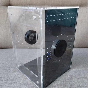 May include: Clear acrylic terrarium with a black base and two black circular vents. The enclosure has a cube shape with a transparent front and sides, and a dark, possibly tinted, back panel. Small holes are visible throughout the structure.