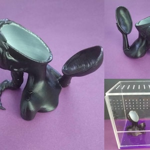 May include: A black 3D printed sculpture of a woman's head and torso with a hollowed-out head and a separate hand holding a mirror. The sculpture is sitting on a purple surface.