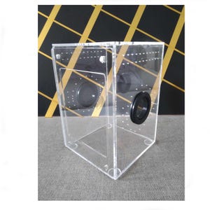 May include: A clear acrylic box with two round black mesh vents. The box is designed for small animals or insects.