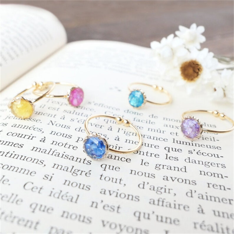 Pode incluir: Seis an&eacute;is de ouro com incrusta&ccedil;&otilde;es de resina de diferentes cores. Os an&eacute;is est&atilde;o sobre um livro com texto. As incrusta&ccedil;&otilde;es de resina s&atilde;o amarelas, cor-de-rosa, azuis, roxas e azul claro.