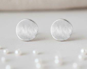 Puede incluir: Un par de pendientes de botón redondos de color plateado. Cada pendiente tiene un acabado de metal cepillado y un diseño simple y minimalista. Los pendientes se exhiben sobre una superficie blanca con pequeñas perlas blancas esparcidas.