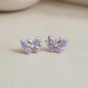 May include: A pair of butterfly-shaped stud earrings featuring light purple enamel wings with silver-toned metal detailing. Each earring is accented with two small, clear, sparkling stones positioned on the upper and center wing sections.