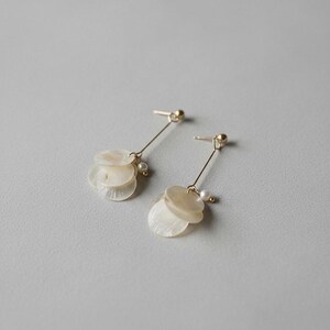 May include: A pair of drop earrings featuring thin gold-toned metal posts with a small ball stud. Each earring suspends a cluster of three translucent, circular, off-white shell discs and a single small white pearl bead attached to the metal wire.
