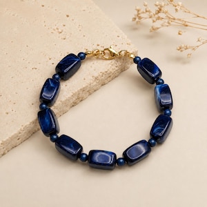 May include: A bracelet featuring rectangular dark blue marbled beads alternating with small round dark blue beads. The piece is finished with a gold-toned metal lobster claw clasp and connecting jump rings, creating a continuous circular design.