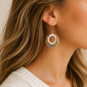May include: A close-up of a colourful beaded hoop earring. The earring features a white inner circle, surrounded by rows of small, multi-coloured beads in shades of yellow, orange, blue, and green. The earring hangs from a silver hook.