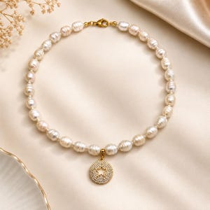 Op de afbeelding: Een parelketting met een gouden sluiting en een hanger. De hanger is een gouden cirkel met een stervormige uitsparing, bezet met kleine, heldere kristallen. De parels zijn gebroken wit en ovaal van vorm.