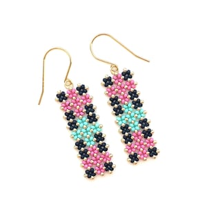 Puede incluir: Un par de pendientes de cuentas con un diseño rectangular. Los pendientes presentan un patrón de cuentas rosas, negras y celeste, con ganchos dorados. Los pendientes se muestran sobre un fondo blanco.