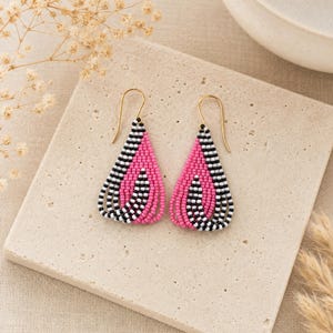 May include: A pair of teardrop-shaped dangle earrings crafted from small seed beads in vibrant pink, black, and white. The beads are arranged in a layered, curved pattern, and each earring features a simple gold-toned metal hook for attachment.
