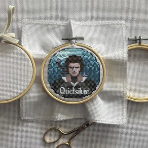 May include: A cross-stitch embroidery of a character named Quicksilver in a wooden hoop. The artwork features a portrait with a blue and teal background. The text "Quicksilver" is stitched below the portrait. Scissors and embroidery hoops are also visible.