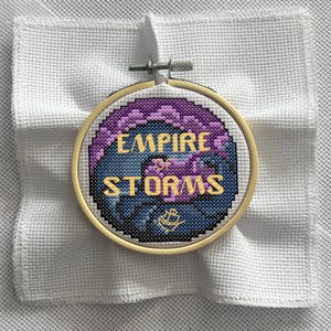 Könnte beinhalten: Ein Kreuzstich-Stickrahmen mit dem goldenen Schriftzug "EMPIRE OF STORMS". Das Design zeigt violette und blaue pixelige Muster und ein kleines Schiffssymbol. Der Rahmen befindet sich vor einem weißen Stoffhintergrund.
