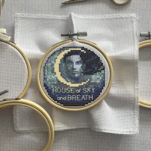 Puede incluir: Bordado de punto de cruz en un aro de madera. El diseño presenta un retrato dentro de una luna creciente, con el texto "HOUSE of SKY and BREATH" en azul claro. El fondo es de tela blanca.