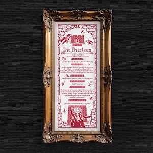 May include: A framed cross-stitch artwork with a gold ornate frame. The artwork features red stitching on a white background, with the text "The Thirteen" and other text and illustrations. The frame is rectangular.