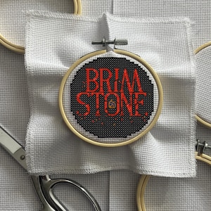 May include: A cross-stitch embroidery hoop with the words "BRIM STONE" in red letters on a black background. The embroidery is set in a wooden hoop and mounted on white fabric. Scissors are visible in the lower left corner.