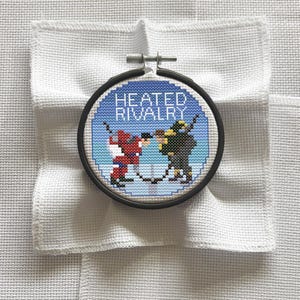 Może przedstawiać: Obręcz do haftu krzyżykowego z motywem hokejowym. Projekt przedstawia dwóch hokeistów w kolorze czerwonym i czarnym, z napisem "HEATED RIVALRY" białymi literami na niebieskim tle. Obręcz umieszczona jest na białej tkaninie.