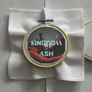 May include: A cross-stitch embroidery hoop featuring the text "KINGDOM OF ASH" in light blue, with a landscape design in shades of black, gray, and orange. The embroidery is set against a white fabric background.