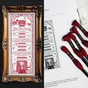 May include: A framed cross-stitch piece with the text "The Thirteen" in red on a white background. The frame is ornate gold. Next to it are red embroidery floss skeins and a printed pattern.