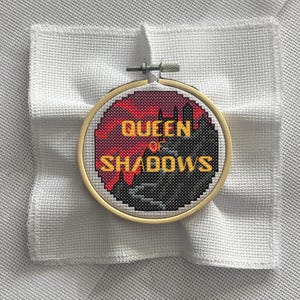 May include: A cross-stitch embroidery in a wooden hoop, featuring the text "QUEEN OF SHADOWS" in yellow, set against a red and black pixelated background. The embroidery is on white fabric.